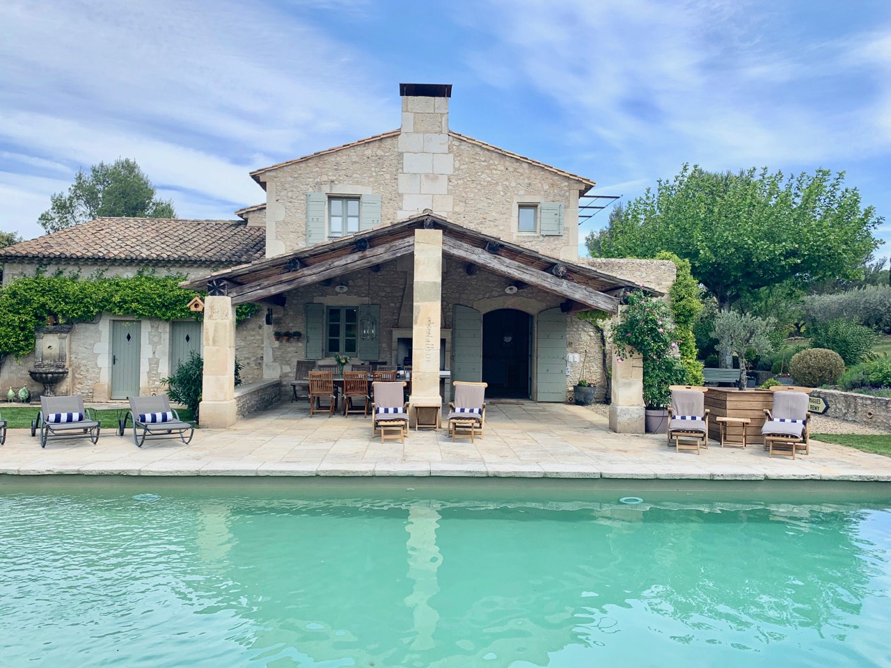 A beautiful stone villa with a covered patio, outdoor seating by a tranquil pool, surrounded by lush greenery under a clear blue sky.